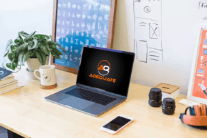 A wooden desk with a laptop displaying the logo "Adequate". Next to it are a smartphone, camera lenses, a plant, a coffee mug, notebooks, and framed artwork. The background features a whiteboard with sketches and diagrams.