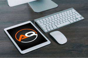 A tablet displaying a logo with the letters "AQ" is placed on a wooden surface next to a white keyboard and a white computer mouse. The tablet's screen shows a black background with an orange and white "AQ" logo in the center.