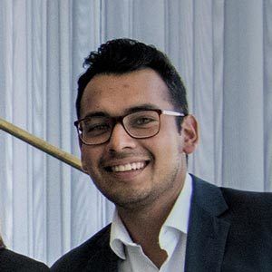 A smiling person wearing glasses and a dark suit with a white shirt stands in front of a light-colored curtain. The person has short dark hair and neatly groomed facial hair.