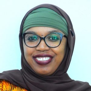 A person wearing a brown hijab, green headscarf, and glasses smiles warmly at the camera. They have dark skin and are wearing a colorful patterned top. The background is light blue, creating a bright and cheerful atmosphere.
