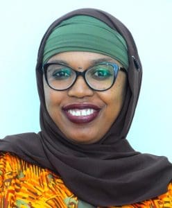 A woman wearing glasses, a green headscarf under a brown hijab, and an orange patterned top smiles at the camera.