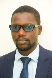 A man with short hair and a beard is wearing dark glasses, a white shirt, a dark blue tie, and a dark blue suit. He is posing against a plain white background, looking directly at the camera with a neutral expression.