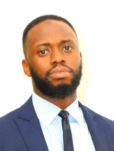 A man with a beard, short hair, and serious expression is dressed in a dark blue suit, white shirt, and black tie. The background is plain and white.