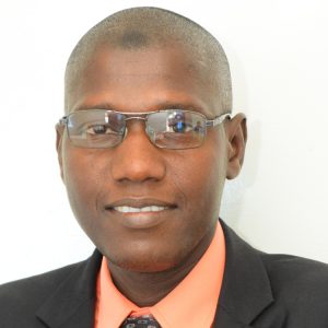 A man wearing glasses, a light orange shirt, and a dark jacket, smiling slightly in front of a plain white background.