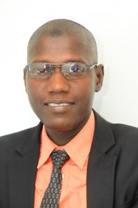 A man wearing glasses, a peach-colored dress shirt, a black suit jacket, and a patterned tie is smiling at the camera. He has a shaved head and is positioned against a plain white background.