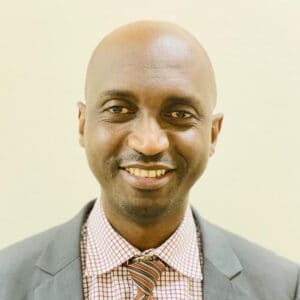 A bald man wearing a gray suit jacket, a red and white checkered shirt, and a striped tie is smiling against a plain light-colored background.
