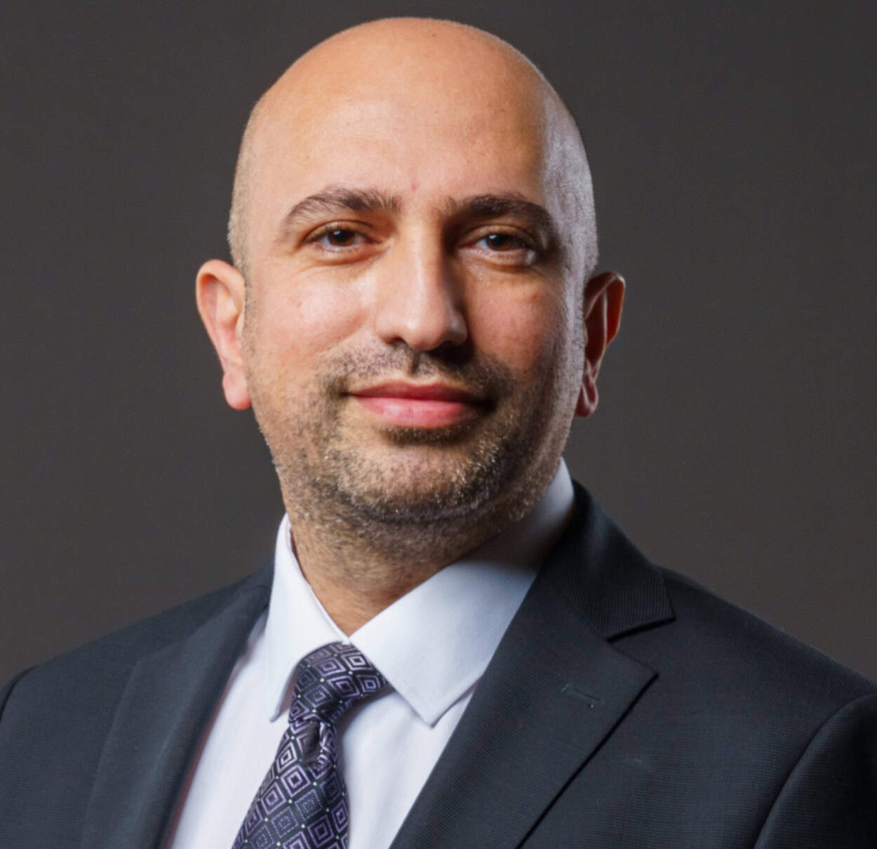 A bald man with light skin and a neatly trimmed beard wears a dark suit, white shirt, and patterned tie, smiling slightly against a plain dark background.