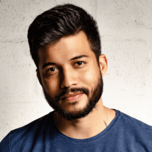 A man with short, dark hair and a beard, wearing a blue shirt, looks directly at the camera with a slight smile. He stands in front of a light-colored, textured wall.