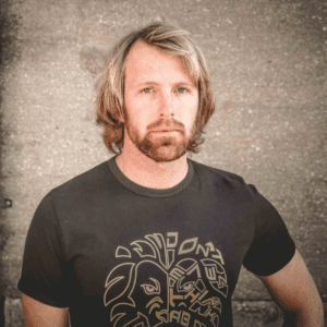 A man with shoulder-length blonde hair and a beard stands against a rough, gray wall. He is wearing a black t-shirt featuring abstract, tribal-style artwork on the front. His expression appears serious.