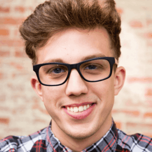 A young person with short, light brown hair and black-framed glasses is smiling while looking at the camera. They are wearing a plaid button-up shirt. The background features a textured wall with exposed bricks.