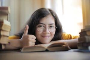 A person with long dark hair and glasses smiles and gives a thumbs-up while leaning over an open book on a table. Stacks of books are visible in the foreground and background, creating a cozy, study-friendly atmosphere.