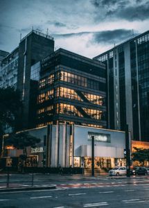 A modern, multi-story Riachuelo store with large glass windows illuminated from within at dusk. The building is surrounded by tall office buildings and a few visible trees. A car is parked on the street in front of the store, with the sky showing early evening hues.
