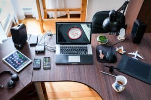 A workspace featuring a laptop with a "Start Engine Stop" screen surrounded by various gadgets, including a tablet, three smartphones, a smartwatch, headphones, a drawing tablet, and a cup of coffee. A small airplane model and a bowl of greenery are also present.