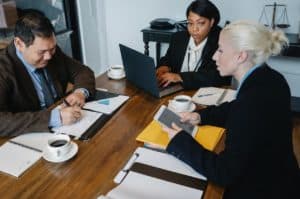 Three colleagues, two women and one man, are seated around a wooden table in a meeting room. They are engaged in discussion, with one woman working on a laptop and the other holding a tablet. The man is writing in a notebook. Coffee cups, papers, and folders are on the table.