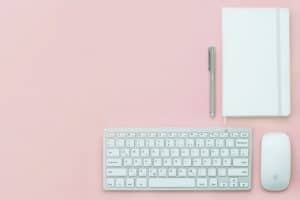 A minimalist workspace on a pink background featuring a white wireless keyboard, a white wireless mouse, an open notebook, and a silver pen placed above the keyboard. The scene conveys a clean and organized setup.
