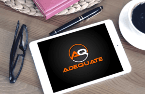 A white tablet on a wooden table displays the logo "Adequate" with the letters "AQ" in a circular design. Nearby are a pair of black eyeglasses, a pen, and an open notebook with a coffee cup partially visible.