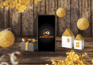 A smartphone displaying the "Adequate" logo stands on a wooden surface, surrounded by small decorative houses with gold roofs, a wrapped gift box with a golden bow, and hanging golden ornaments and flowers in the foreground.