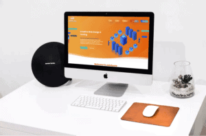 A sleek, modern workspace features an iMac displaying a colorful website homepage for "Creative Web Design & Hosting." On the white desk are a round Harman Kardon speaker, a white keyboard, a white mouse on an orange pad, and a glass terrarium.
