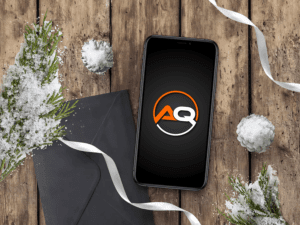 A smartphone displaying an "AQ" logo on a rustic wooden surface, surrounded by snow-covered pine branches and decorative snowballs. A black envelope with a white ribbon rests nearby, adding a festive and wintry touch to the scene.