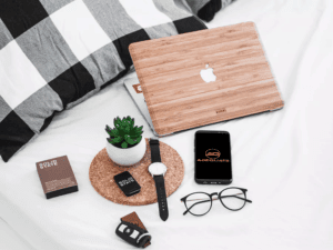 A neatly arranged workspace on a white bed features a wooden laptop, a black smartphone with "ADEQUATE" on the screen, eyeglasses, a wristwatch, a small potted plant, a leather keychain, a solid state gift box, and a small solid state item.