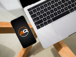 A smartphone displaying a logo with the letters "AQ" in orange and white on a black background is placed next to a silver laptop. The devices are on a light-colored table with wooden legs. The laptop's keyboard is visible, showing a touch bar and trackpad.