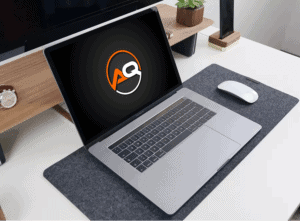 A sleek laptop with a black screen displaying a logo with the letters "AQ" in white and orange. The laptop is placed on a dark grey desk mat, accompanied by a white wireless mouse. The background features a wooden stand with a potted plant and other small items.