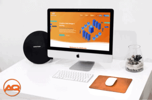 A sleek desktop setup featuring an Apple iMac displaying a colorful web design and hosting website. The desk also includes a round Harman Kardon speaker, a mouse on a brown mousepad, and a small plant in a pot, all on a clean white surface.