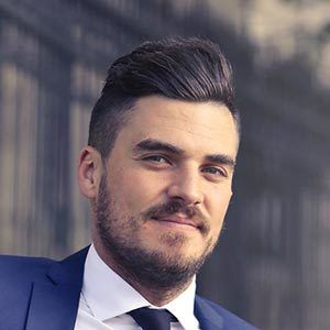 A man with short dark hair and a beard, wearing a blue suit jacket, white shirt, and black tie, smiles slightly. The background appears to be an outdoor setting with a blurred view of a building or structure.