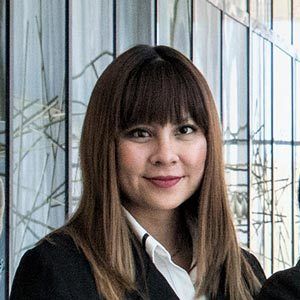 A woman with long, straight, brown hair and bangs is standing indoors. She is wearing a dark blazer over a white shirt and is smiling at the camera. The background features a glass wall with a modern, minimalist design.
