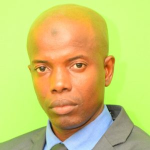 A bald man wearing a dark suit, blue shirt, and black tie poses in front of a bright green background. He has a serious expression.