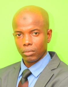 A man with a serious expression, a bald head, and wearing a dark suit, blue shirt, and black tie, is posing in front of a bright green background.