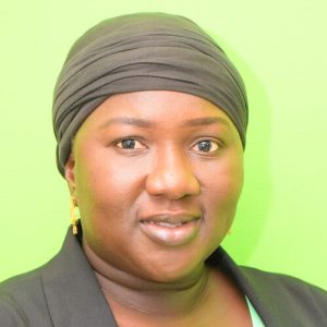 A person wearing a black headscarf and black blazer is staring directly at the camera. The background is a bright green color. The person is smiling subtly, with gold earrings visible.