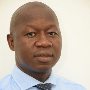 A man with a shaved head wearing a light blue dress shirt and a navy blue tie with a dotted pattern stands against a plain white background. He is looking directly at the camera with a neutral expression.