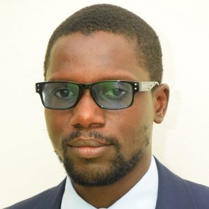 A man with short black hair, a beard, and glasses is wearing a dark suit jacket, a white shirt, and a tie. He has a neutral expression and is looking directly at the camera. The background is plain white.
