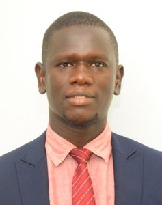A man with short hair, wearing a dark blue suit jacket, light red dress shirt, and a red striped tie, posed against a plain white background.