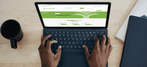 Top-down view of a person’s hands typing on a laptop keyboard. The laptop screen displays a webpage for "Agib Bank Ltd" with application forms for individual account opening. A black coffee mug, a notepad, and a pen are placed on the desk beside the laptop.