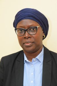 A woman wearing a dark blue headscarf, black glasses, and a black blazer over a light blue shirt. She has a calm expression and is looking directly at the camera. The background is a plain, light-colored wall.