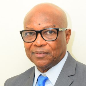 A bald man wearing glasses, a gray suit, a blue shirt, and a blue tie stands against a plain, light-colored background. He is looking directly at the camera with a neutral expression.