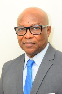 A man with a bald head and wearing glasses is dressed in a gray suit, light blue shirt, and blue tie. He is looking at the camera with a neutral expression. The background is plain and light-colored.