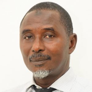 A middle-aged man with a short black and grey beard, wearing a light-colored checkered shirt and a dark tie, looks directly at the camera against a plain white background. His expression is calm and neutral.