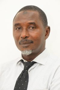 A man with short hair and a salt-and-pepper goatee, wearing a light-colored striped shirt and a dark polka-dotted tie, looks directly at the camera against a plain white background.